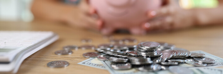 Banknotes and coins on table and blurry woman with piggy bank in background.