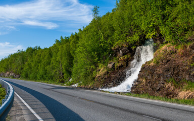 Waterfall flows from green forest just next to the asphalt road