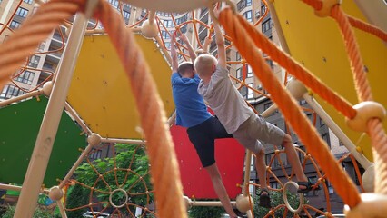 Two active boys climb a rope slide on the playground in the courtyard, they hang on their hands and pass various tests.