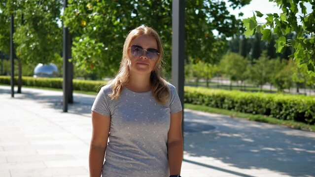 A young woman with blonde hair wearing sunglasses walks through a modern city park in summer in sunny hot weather. The girl walks slowly and looks at the camera.