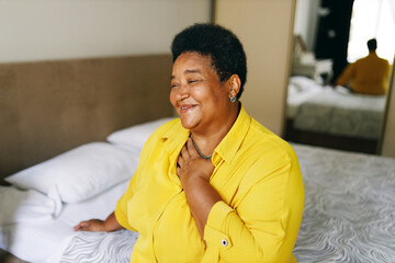 Happy cheerful grateful african american grandmother sitting on bed in yellow shirt keeping hand on chest smiling, being thankful for best life, thinking about her past, funny unforgettable moments