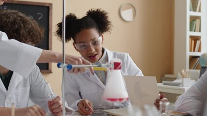 Waist up of preteen African American schoolgirl wearing safety glasses and white lab coat conducting chemical experiment during Science lesson with teacher and multiracial classmates - Powered by Adobe