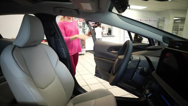 Pretty blonde mature woman looking in driver side door window and then opening door and getting in drivers seat considering buying from a dealership.