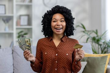 Portrait of a young African American woman sitting on the sofa at home, holding a credit card and cash money, looking enthusiastically and smiling at the camera