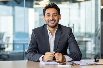 Portrait of young hispanic businessman inside office, boss in business suit smiling and looking at camera, experienced satisfied man at workplace at desk.