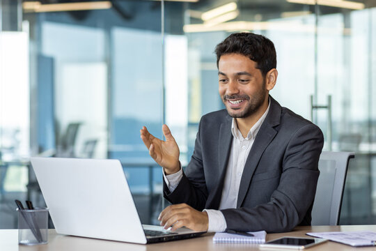 Young successful arab businessman inside office at workplace talking remotely with colleagues and partners, man using laptop for video call, satisfied boss smiling and gesturing with hands joyfully.