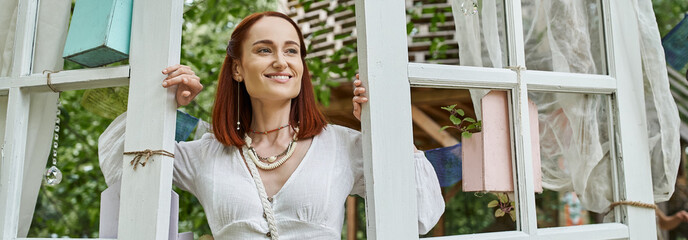 joyful and stylish redhead woman standing near doors outdoors in retreat center, banner