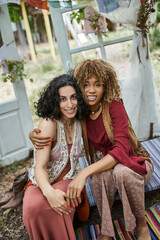 smiling interracial girlfriends in boho outfits hugging and looking at camera in retreat center