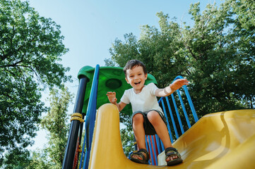 happy preschooler boy playing on slide on playground in summer
