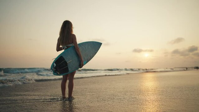 Tourist woman surfer in swimsuit with surfboard stands on ocean sandy beach at sunset admires waves. Summer sport activity on tropics resort. Sporty slim girl. Extreme water sport surfing on vacation.
