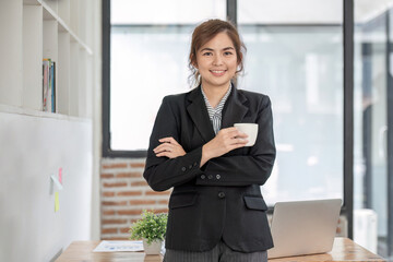 Portrait, Confident smiling asian businesswoman in suit standing in office with arms folded and holding coffee cup.