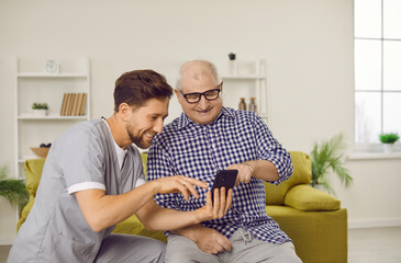 Happy elderly man using mobile phone. Carer helps old man use smartphone. Worker looks at digital...