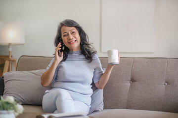 A happy 60s Asian woman enjoys her coffee and talking on the phone