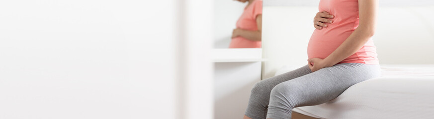 Asian pregnant woman sitting embracing her belly on bed at home. Banner.