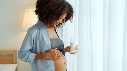 Portrait of Black African American Pregnant woman embracing her tummy and holding a glass of milk to drink. Expecting mother at home. Pregnancy. Motherhood.
