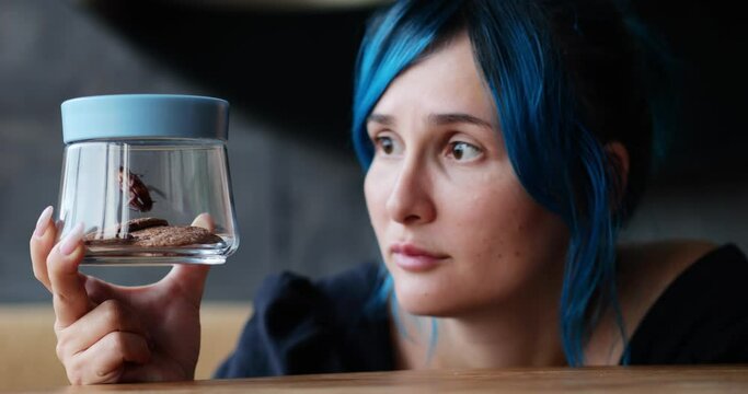 Young woman caught or found a large cockroach in glass jar with cookies, holds it in her hand and looks at the insect with disgust and curiosity. Close up shot with shallow focus