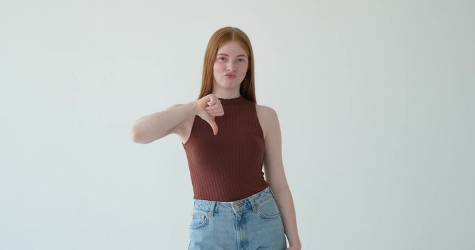 A dissatisfied red haired girl is indicating a thumbs-down gesture on a white background. Her discontent is evident from her facial expression and the downward gesture of her thumb.