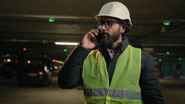 Professional industrial manufacture business industry worker Arabian bearded engineer Indian man in glasses builder in safety uniform hard hat talking phone speak smartphone at parking lot at factory