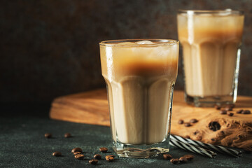 Ice coffee in a tall glass with cream poured over, ice cubes and beans on a dark concrete table.