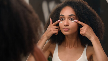 Ethnic girl beauty routine in morning young African American woman applying face cream under eyes anti-aging skin care dark eye circles moisturizing organic natural cosmetic facial mask looking mirror