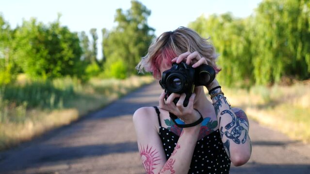 Happy female hipster with pink hair taking photos on photocamera at empty country road. Carefree punk girl with tattoos posing on camera at rural driveway. Hippie woman doing pictures outdoor