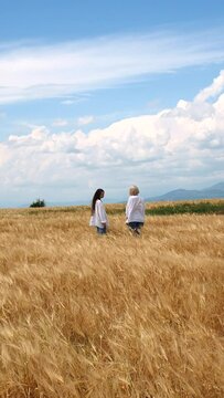 Two girls in white linen shirts and jeans walking in a rye field on a windy day
