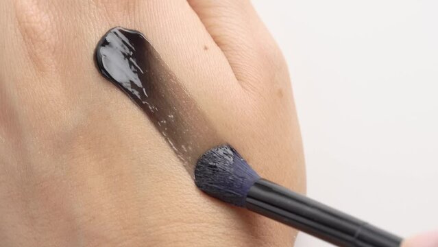 Clay or charcoal mud cream in jar on white background. Brush gains black cream mask applying on hand skin. Swatch black cream mack for face