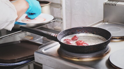 Chef in a restaurant kitchen fry chicken liver in hot pan. Professional woman cook in blue gloves puts red fresh meat on the hot surface of the pan. Preparing ingredients, health dietary food delivery
