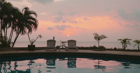 Woman relaxes sitting near pool on tropical island hotel resort. Bright colorful sunset sky and coconut palms reflected in water. Travel, tourism, holiday spa vacation. Slow motion portrait zooms in © Anastasia Pro