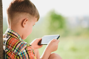 The child looks into the phone while sitting in the summer on the street. Little boy with a mobile in his hands.