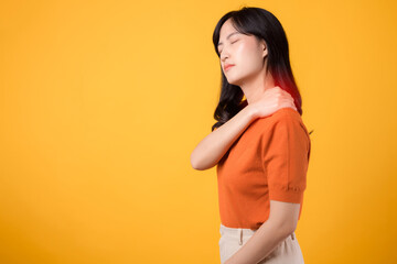 Fototapeta na wymiar Vigilant young Asian woman in her 30s, wearing an orange shirt, holds her pain shoulder on yellow background. Neck ache therapy medical office syndrome concept.