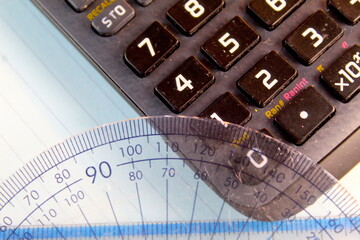 A photo of a mathematics protractor on top of a black calculator on a white desk. 