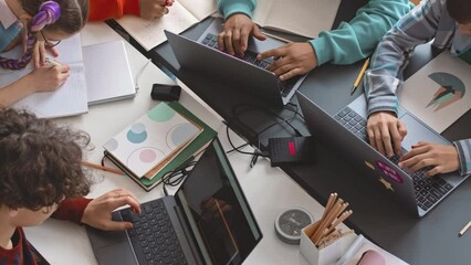 Top view of diverse preteen school children using laptops and taking notes in their copybooks while studying programming together at desk in classroom - Powered by Adobe
