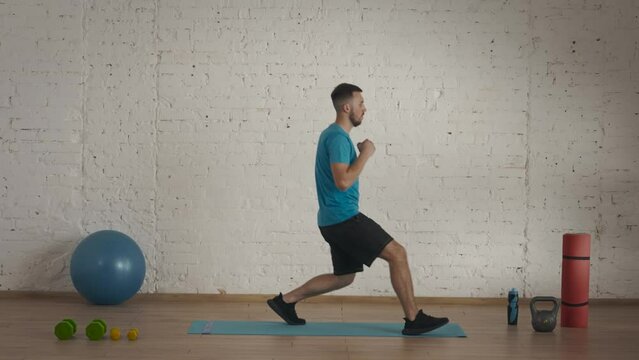 Man fitness instructor in sportswear doing lunges exercise for online class at the home studio. Side view.