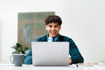 Happy european freelancer man working on laptop wearing headphones on neck and smiling, sitting in...