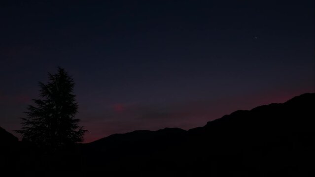 Time lapse of the moon over a hill. View of a night to day sky with the silhouette of a fir tree on a mountain.