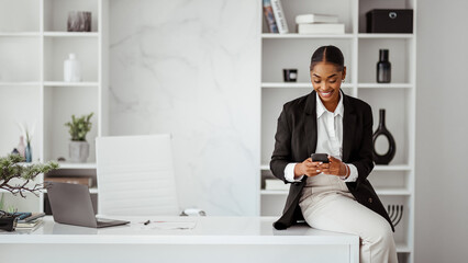 Successful black businesswoman in formal wear sitting leaning at desk with laptop and using phone...