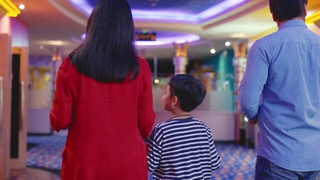 Back of family with Caucasian father, Asian mother and childs walk together in front of cinema theater and look for their hall for them to enjoy to watch the movie.