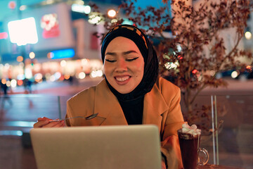 A hijab woman enjoying a sweet dessert at a coffe while working on her laptop during the nighttime in a bustling urban setting.