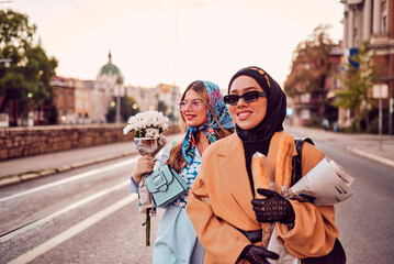 Couple woman one wearing a hijab and a modern yet traditional dress, and the other in a blue dress and scarf, walking together through the city at sunset. One carries a bouquet and bread, while the