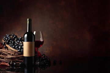 Glass and bottle of red wine with blue grapes on a black reflective background.