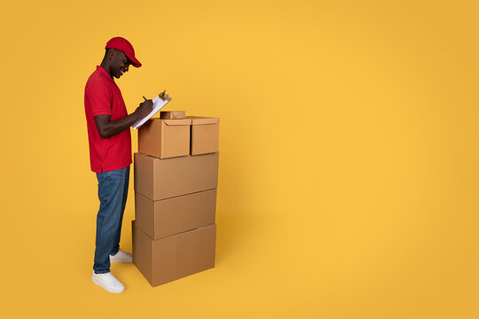 Cheerful millennial african american guy courier in uniform making notes in warehouse, cheks many boxes