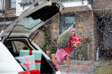 Woman in red sweater and hat carries wrapped Christmas tree and gift box in porch of her house during a snow fall. Concept of preparing for winter holidays