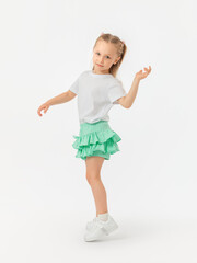 A beautiful 5-year-old girl is dancing on her toes in a skirt, T-shirt and sneakers on a white background. Copy Space