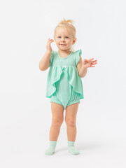 A blonde girl of 2 years old smiles, stands and holds her hand to her face, straightens her hair on a white background in a green bodysuit dress and socks.