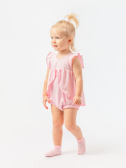 A cute 2-year-old toddler girl with blue eyes in a pink dress walks in socks on a white background.