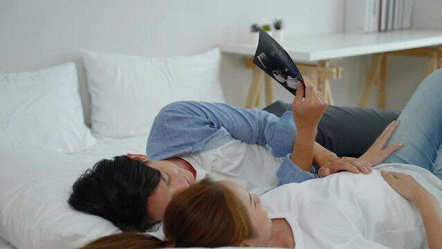 Asian couple lying down looking at the ultrasound results of their baby in the bedroom, concept of initiation, family relationship
