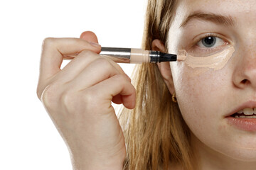 young blond woman apply concealer under the eye on a white background