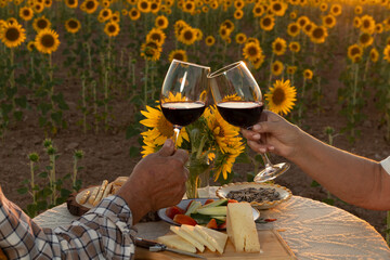 hands of senior couple toasting with glasses of wine having dinner al fresco in sunflower fields
