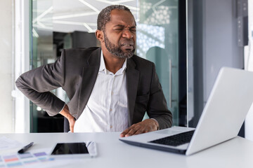 Obraz premium Sick mature african american businessman having severe back pain, man in business suit at workplace sitting job overworked and overtired.
